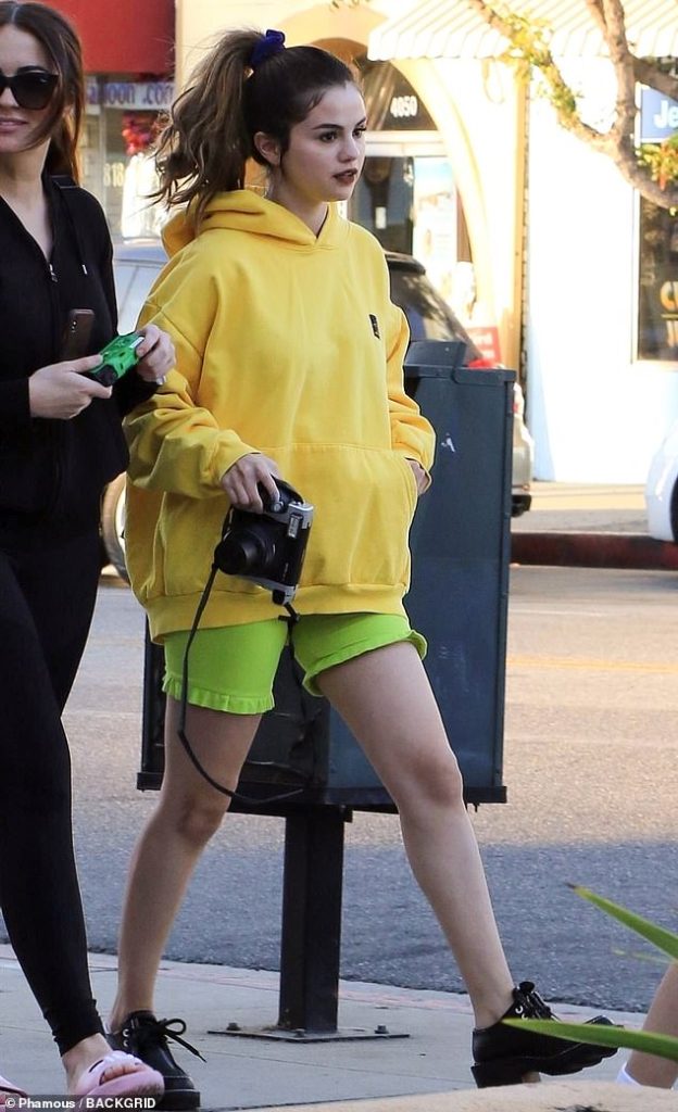 Selena Gomez Rocks a Vibrant Ensemble with Neon Sweatshirt and Biker Shorts, Capturing Moments with an Instant Camera